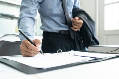 Homem veste roupa social e segura blazer em uma das mãos. A imagem mostra apenas parte de seu coro que está debruçado sobre a mesa, assinando um papel. Representando a assinatura recorrente.