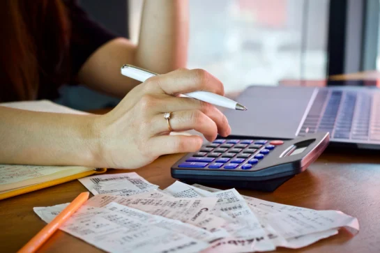 Mão feminina segurando um lápis enquanto digita na calculadora. Ela está fazendo contas, que estão a sua frente, organizando a carteira de cobrança de seus clientes.