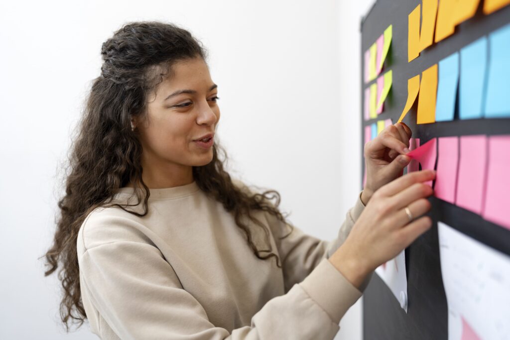 Mulher está em seu escritório, colocando post its em um mural. A imagem representa como organizar uma agenda de trabalho.