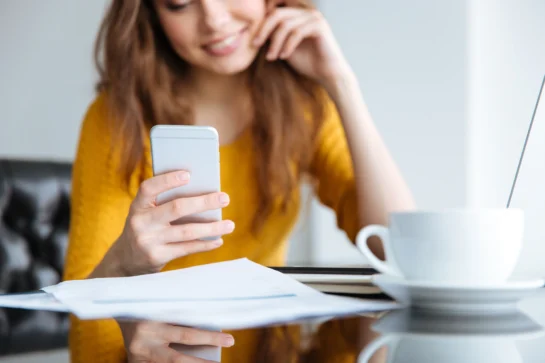 Mulher sorrindo enquanto olha para um boleto por SMS recebido no celular, com boletos sobre a mesa e uma xícara de café.