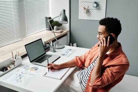 Empreendedor sentado em frente a mesa, enquanto fala no telefone. Ele está aprendendo como organizar agenda de trabalho, com instruções de outra pessoa.