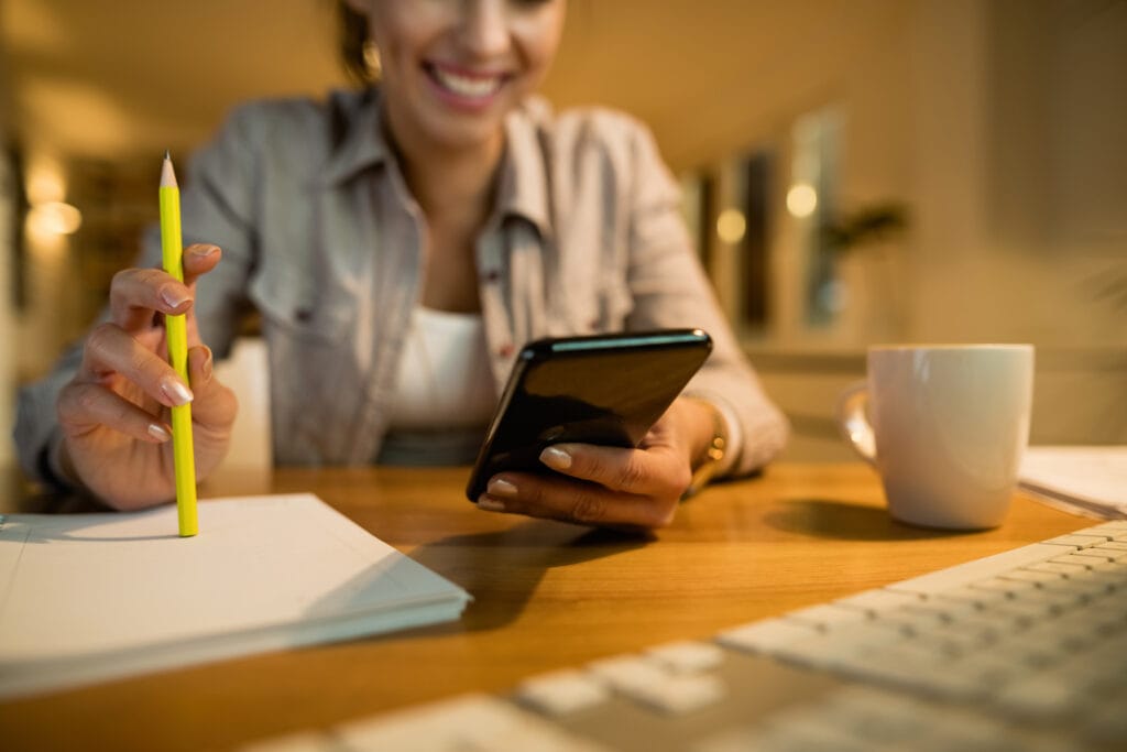 A imagem mostra uma mulher mexendo no seu celular, com o computador e seu café sobre a mesa. Ela está programando o envio de boleto por sms.