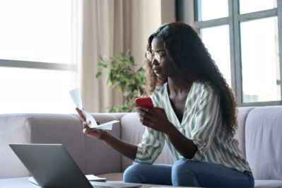 Mulher segurando um celular e um papel, ao lado de um notebook, verificando lembrete de pagamento em casa.