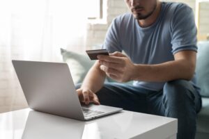 Na imagem, temos um homem na frente de um computador. Ele está segurando um cartão de crédito, finalizando uma compra online. Isso representa como vender produtos na internet.