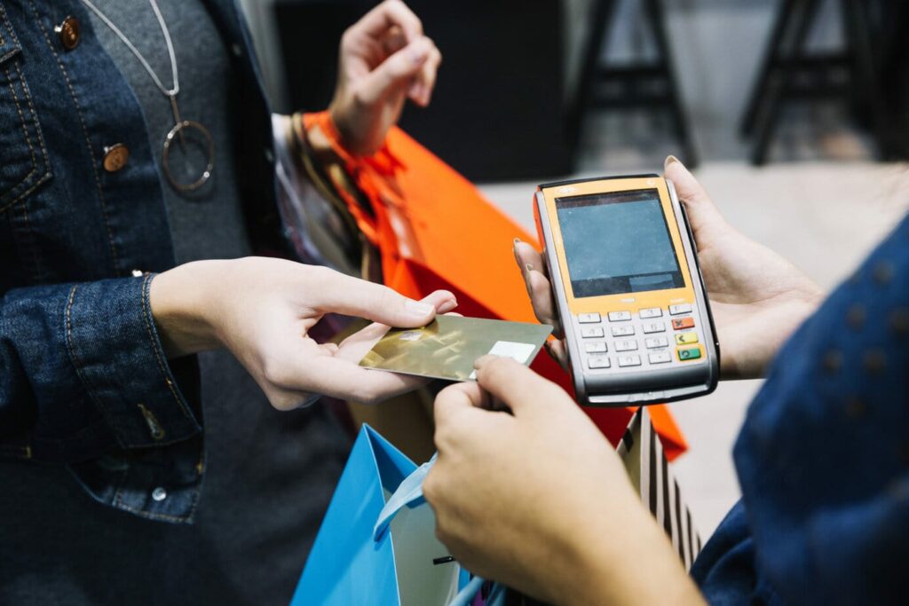 Na imagem, há uma cliente com diversas sacolas de compras no braço. Na frente dela, temos uma atendente segurando um cartão e uma máquina de pagamento. Isso representa como aumentar as vendas no varejo.