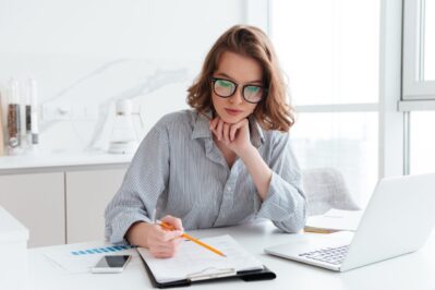 A imagem mostra uma mulher. Em sua mesa, há um notebook aberto e alguns gráficos impressos. Ela está pesquisando como organizar as finanças da empresa.