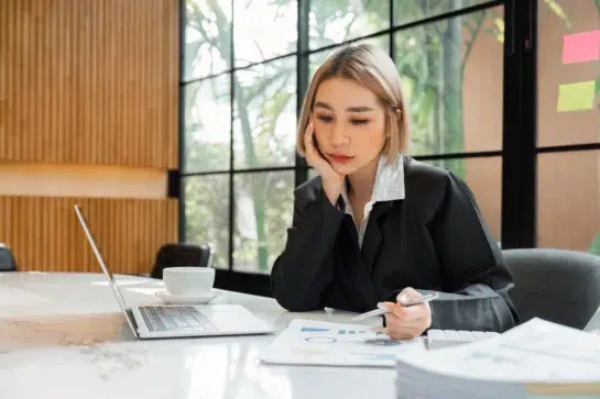 Empresária lê gráficos em mesa, com um computador em sua frente. Ela está estudando sobre a possibilidade de emitir nota fiscal automaticamente.