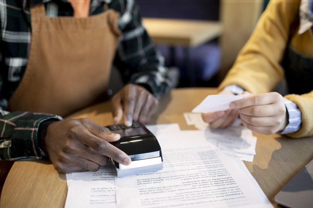 Na imagem, temos um empreendedor e seu funcionário no local de trabalho. Um deles está segurando uma maquina de cartão e o outro está com alguns papeis em mãos. Eles estão entendendo as diferenças entre os tipos de notas fiscais.