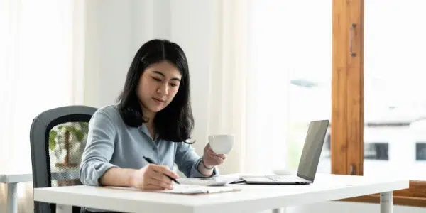 Mulher tomando café enquanto faz anotações em um escritório, com laptop aberto checando a sua conta MEI.