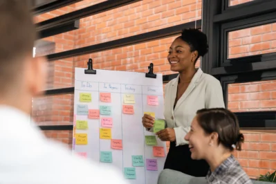 Mulher apresenta quadro de tarefas em equipe, explicando de forma prática como funcionam as vantagens e desvantagens do Scrum.