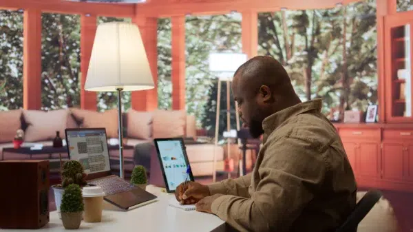 Homem trabalhando, escrevendo em papel, com computador e tablet em cima da mesa. Ele está conferindo os dados para emissão de nota fiscal.