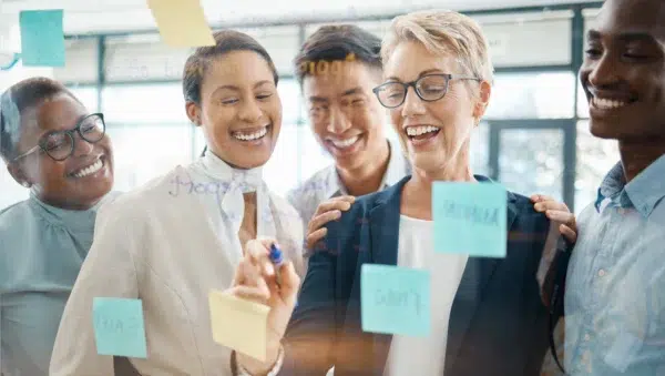 Pessoas sorrindo e escrevendo em um quadro transparente, com post-its. Estão escrevendo o plano de ação para motivar equipes.