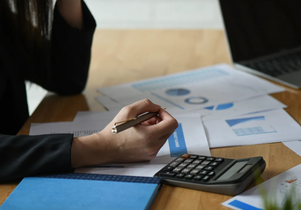 Mulher segurando caneta debruçada sobre documentos contábeis com uma calculadora em cima da mesa, realizando a contabilidade para empresas.