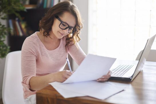 Na imagem, temos uma microempreendedora em seu escritório, mexendo no seu computador com um boleto em mãos. Ela está pesquisando formas de como protestar um boleto.