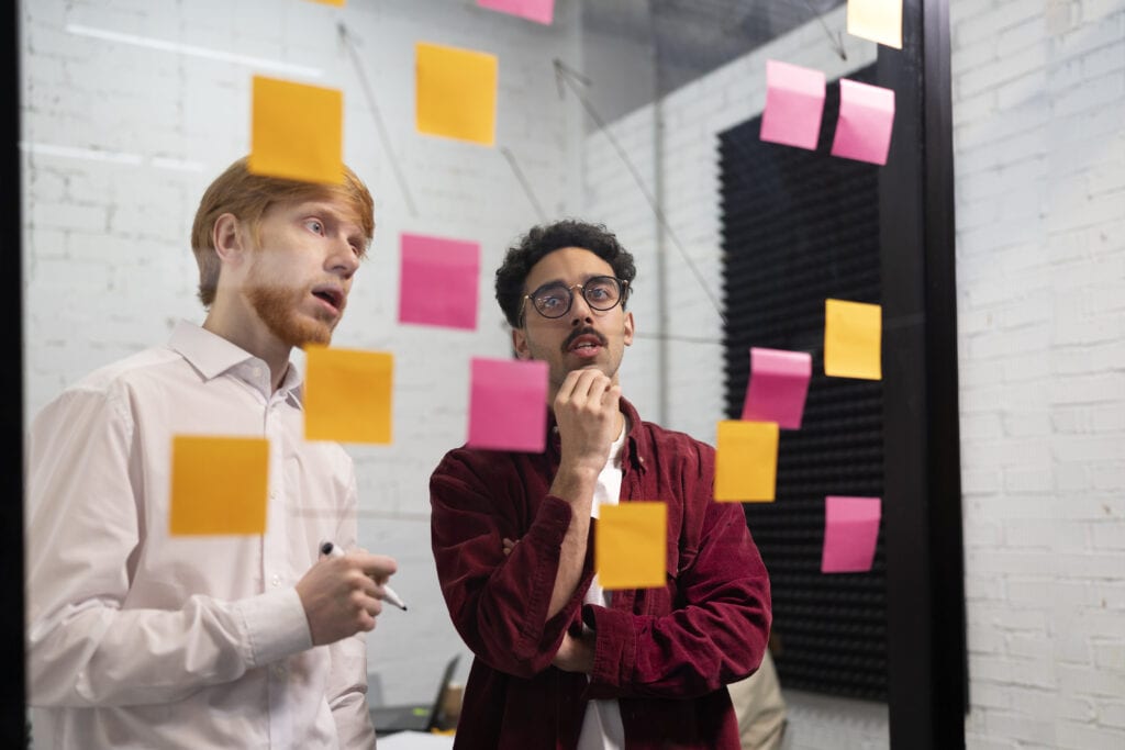 A imagem mostra dois lideres de uma empresa em um escritório, na frente de uma parede de vidro com vários papéis adesivos. Eles estão construindo o planejamento estratégico empresarial.
