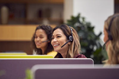 Mulher sorri com fone de ouvido. Ela trabalha com atendimento ao cliente e está na empresa, com pessoas ao redor.