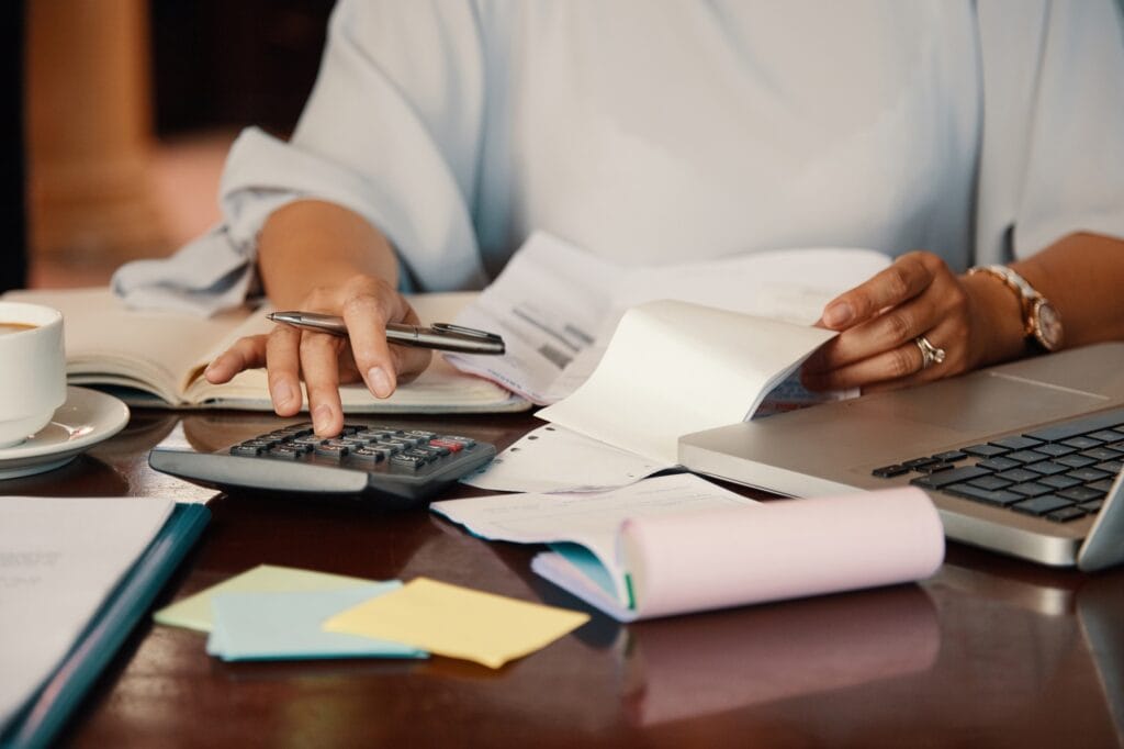 Mãos sobre uma mesa com notebook, papéis e calculadora. A imagem busca representar uma pessoa calculando a tarifa de boleto bancário.