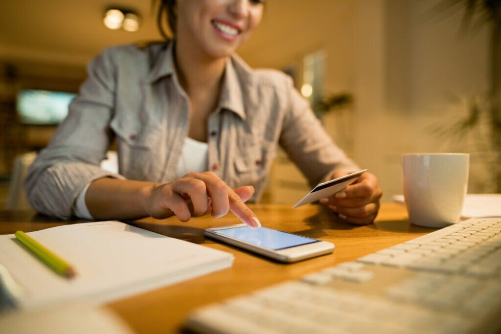 A imagem mostra uma empreendedora em seu escritório, com um cartão de crédito em uma das mãos. Sobre a mesa estão um celular e um caderno, utilizados para fazer a gestão de cobrança.