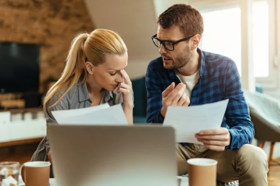 Casal de empreendedores sentados em frente a dois computadores. Estão tentando aprender como reduzir custos na empresa enquanto conversam.