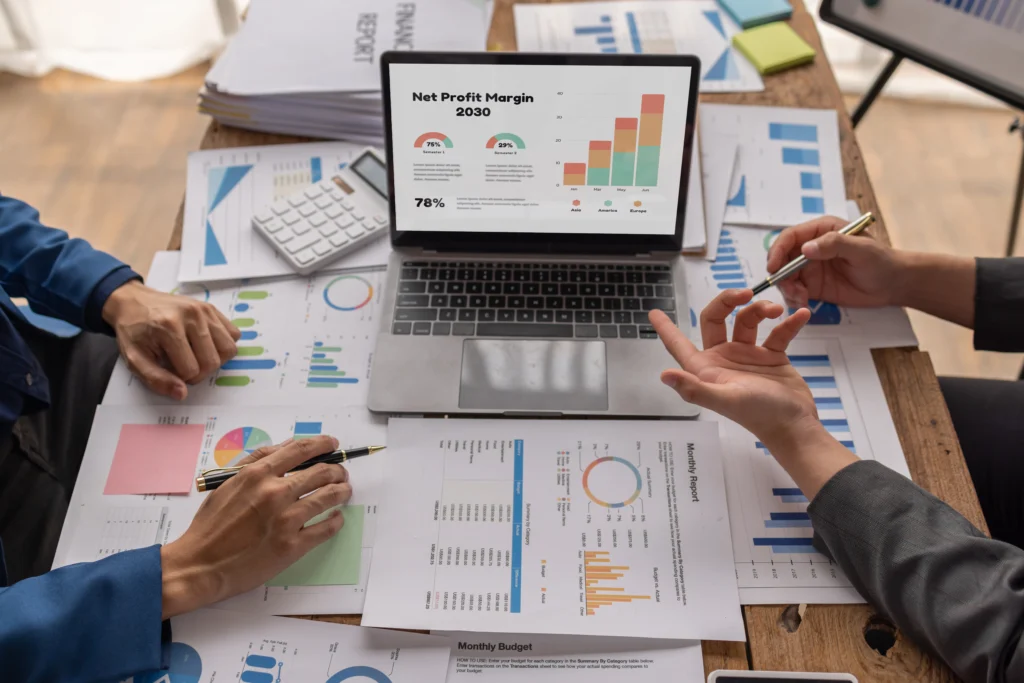 Imagem mostra duas pessoas apoiadas em uma mesa com notebook mostrando gráficos e mesa cheia de papéis com gráficos. Eles estão fazendo o controle de contas a receber.