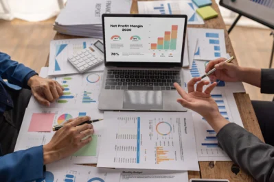 Imagem mostra duas pessoas apoiadas em uma mesa com notebook mostrando gráficos e mesa cheia de papéis com gráficos. Eles estão fazendo o controle de contas a receber.