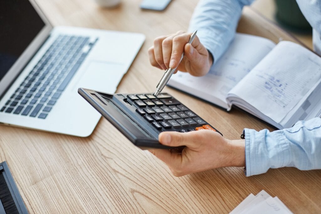 Na imagem, temos um empreendedor em seu escritório, de frente para o notebook. Ele está com uma calculadora, fazendo o planejamento financeiro empresarial da sua empresa.