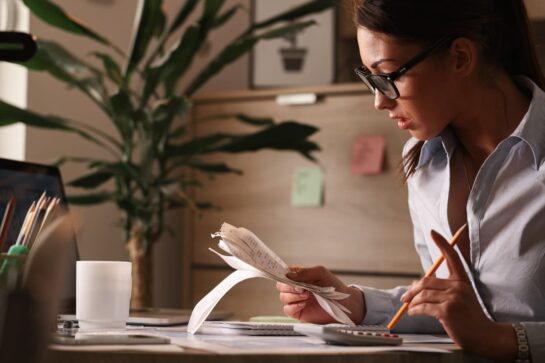 A imagem mostra uma microempreendedora no escritório, em frente ao computador, segurando alguns recibos e uma calculadora. Ela está fazendo a gestão de contas de sua empresa.