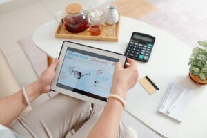 Na imagem, tem uma pessoa sentada segurando um tablet nas mãos, na tela tem uma loja virtual. Em sua frente tem uma mesa, nela tem uma calculadora e um cartão de crédito, representando a venda a prazo.