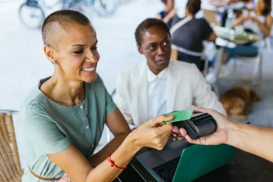 Mulher sorridente realiza uma venda a prazo com cartão, aproximando-o de uma maquininha em um café ao ar livre.