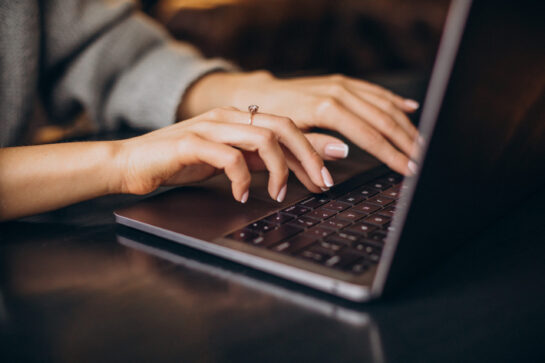 Mulher está no escritório, sentada em frente a uma mesa, mexendo no computador. Ela está escrevendo uma carta de cobrança para seus clientes.