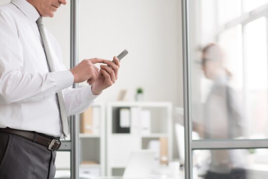 Homem usa roupa social e está em pé segurando um celular nas mãos. A imagem busca representar o uso de um sistema de cobrança automatizado pelo celular.