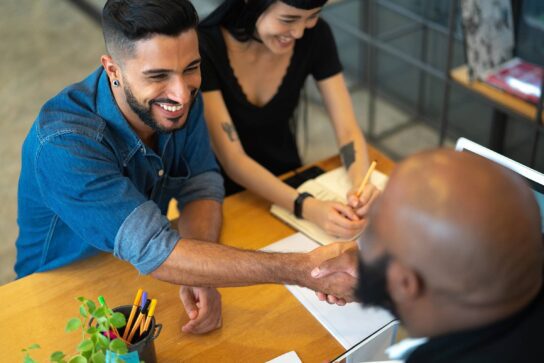 A satisfação do cliente está representada na imagem. No cenário há uma mesa com algumas folhas encima. Deum lado da mesa há um casal, o homem tem cabelos pretos e barba e está sorrindo, ele veste uma blusa jeans. A mulher, tem cabelos pretos e veste um vestido preto, ela está sorrindo. Ambos apertam as mãos de um homem que está do lado oposto da mesa. Ele é careca e usa óculos e veste um terno preto.