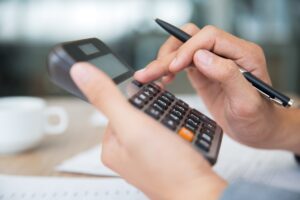 A imagem mostra um empresário em seu escritório, com uma calculadora nas mãos. Ele está aprendendo como calcular a amortização das dívidas de sua empresa.