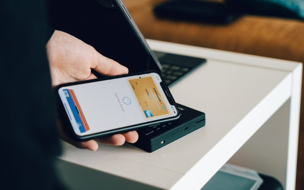 Mão segura o celular e aponta para uma mesa. Na mesa tem um notebook. A imagem busca representar o que é gateway de pagamento.
