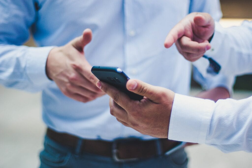 A imagem as mãos de dois homens, um deles segura o celular em uma das mãos e a outra aponta para o aparelho, representando o controle de pagamentos de clientes.