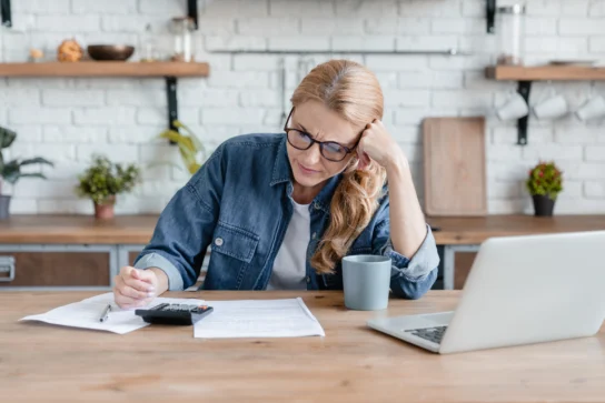 Mulher sentada à mesa fazendo contas, com papéis, calculadora e computador em sua frente. Ela estuda programa para emitir nota fiscal.