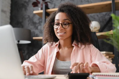 Mulher empreendedora sentada à mesa, em frente a um computador, mexendo em calculadora. Está aprendendo a criar a régua de cobrança de sua empresa.