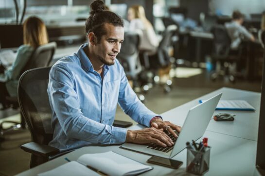 Homem está sentado frente a uma mesa. Na mesa tem um notebook, um celular, um pote com canetas e um caderno. O homem veste uma blusa azul e está com as mãos sobre o notebook. A imagem busca representar como usar um sistema de cobrança.