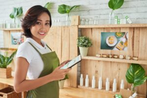 Mulher vestindo blusa branca e avental verde, segura um celular nas mãos. O cenário é uma loja de plantas, com prateleiras de madeira. A imagem busca representar a consulta serasa para empresas.