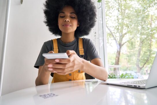 Mulher negra está buscando como funciona o pix. Com braço apoiado em cima da mesa, ela segura o celular nas mãos para ler o QR code sobre a mesa.