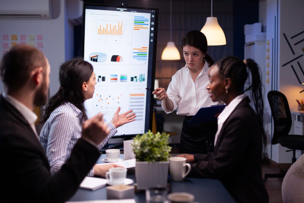 A imagem mostra uma equipe conversando sobre o fluxo de caixa da empresa num escritório, com a tv mostrando os números do controle. Eles estão fazendo a gestão financeira empresarial.