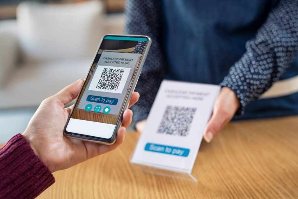 Duas mãos, de pessoas diferentes, uma segura um celular nas mãos e a outra um totem de mesa com um QR Code. A imagem busca representar o Pix para empresas.