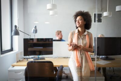 Mulher negra, está em pé em frente a mesa do escritório. O ambiente tem um computador, estante, luminária e cadeira. A imagem busca representar Como funciona o Pix Banco do Brasil.