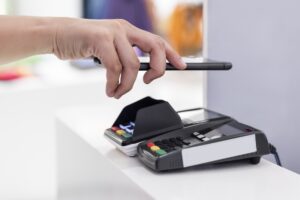 Uma mão segura um celular, apontando para uma máquina de cartão que está sobre um balcão. A imagem representa como funciona o Pix bradesco para pagamento.