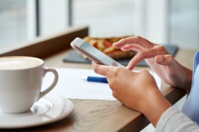 Mãos segurando um celular, frente a uma mesa. Encima da mesa tem um píres com uma xícara e uma folha de papel branca. A imagem busca representar se o Pix cobra taxa.