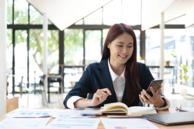 Mulher sorrindo, usando celular em escritório com documentos e gráficos, verificando se o Pix cobra taxa para transações.