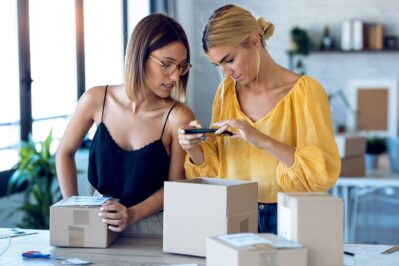 Duas mulheres estão em uma loja. Uma veste uma blusa amarela e a outra uma blusa preta. A de blusa amarela segura o celular e aponta para uma caixa. Ambas estão olhando para a tela do aparelho. A imagem busca mostrar como funciona o Pix Santander.