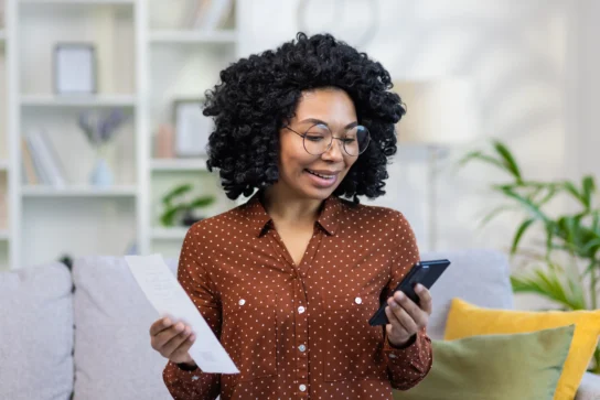 Mulher sorridente usa um aplicativo de cobrança no celular enquanto segura um boleto, sentada em um sofá com almofadas coloridas.