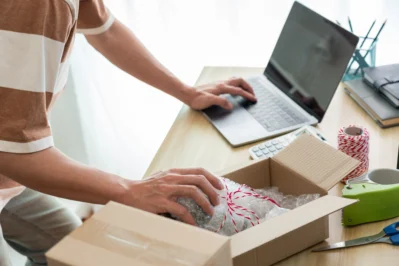 Homem mexendo no computador e em uma caixa de um produto para entrega. Está calculando a precificação do produto para envio.