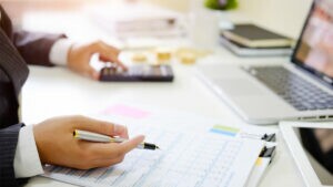 Mãos femininas seguram caneta e mexem em calculadora, com o objetivo de identificar as despesas fixas e variáveis do negócio. Ao fundo, há um notebook e materiais de escritório.